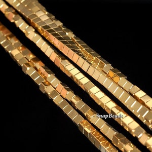 Könnte beinhalten: Goldfarbene quadratische Perlen, die zu einer langen Schnur aufgefädelt sind. Die Perlen sind versetzt angeordnet und erzeugen so eine strukturierte Optik. Die Schnur liegt auf einer schwarzen Oberfläche.