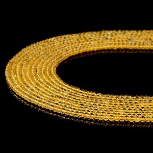 Pode incluir: Close-up de vários fios de contas de pedras preciosas amarelo dourado. As contas, pequenas e redondas, são de tamanho uniforme, criando uma linha suave e curva sobre um fundo preto. As contas refletem a luz, dando-lhes uma aparência brilhante.