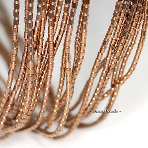 May include: A close-up of a strand of copper-colored metal beads. The beads are small and cylindrical, with a slightly rounded shape. The beads are strung together on a thin wire, and the strand is coiled into a loose spiral.