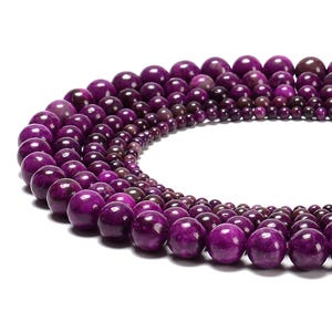 May include: Close-up of a strand of purple gemstone beads. The beads vary in size, with the largest ones at the front. The color ranges from deep purple to lighter shades, creating a gradient effect. The beads are round and polished, reflecting light.