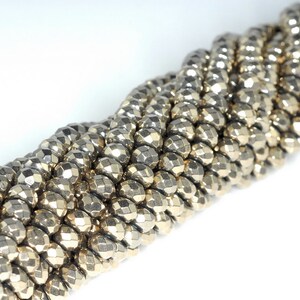 May include: Close-up of a strand of faceted, metallic beads. The beads are a muted gold color and have a geometric, multi-faceted design. The beads are strung together, creating a textured, rope-like appearance. The background is white.