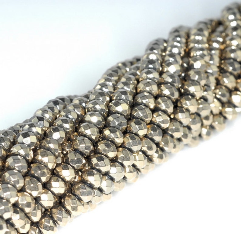 May include: Close-up of a strand of faceted, metallic beads. The beads are a muted gold color and have a geometric, multi-faceted design. The beads are strung together, creating a textured, rope-like appearance. The background is white.