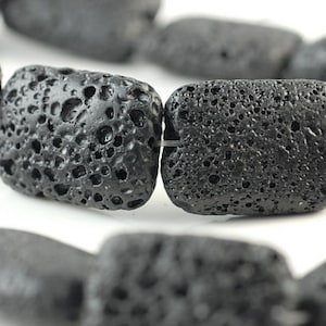 Puede incluir: Una hebra de cuentas de roca volcánica negra, cada cuenta es rectangular y tiene una textura rugosa y porosa.