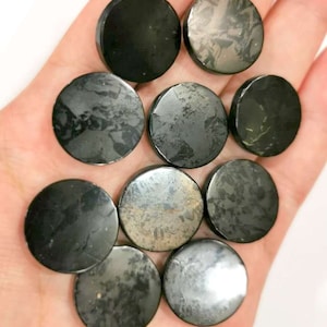 Puede incluir: Diez piedras negras y circulares con una superficie pulida y un patrón moteado plateado. Las piedras se sostienen en una mano. Las piedras miden aproximadamente 2,5 cm de diámetro. El fondo está desenfocado.