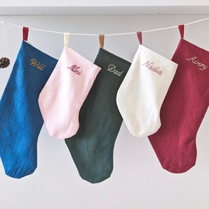 Puede incluir: Cinco calcetines de Navidad personalizados colgados de un estante blanco. Los calcetines son azules, rosas, verdes, blancos y rojos. Los nombres "Will", "Mlei", "Dad", "Nadia" y "Avery" están bordados en los calcetines.