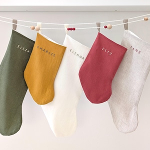 Op de afbeelding: Vijf kerstsokken van olijfgroen, mosterdgeel, wit, rood en grijs linnen hangen aan een touwtje met houten kralen. Elke sok is geborduurd met een naam: Eliza, Charlie, Elinor, Fitz en Jane.
