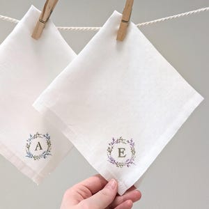 Könnte beinhalten: Zwei weiße Leinen-Taschentücher, eines an einer Wäscheleine mit einer Holzklammer aufgehängt, das andere von einer Hand gehalten. Jedes Taschentuch hat ein florales Kranzdesign mit einem Buchstaben in der Mitte. Der Buchstabe "A" ist in einem blau-grünen Kranz, und der Buchstabe "E" ist in einem lila-grünen Kranz.