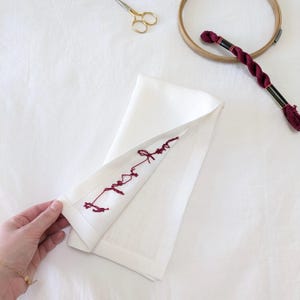 May include: A white linen napkin with a red embroidered design. The embroidery is a simple, free-flowing design that looks like a handwritten word.