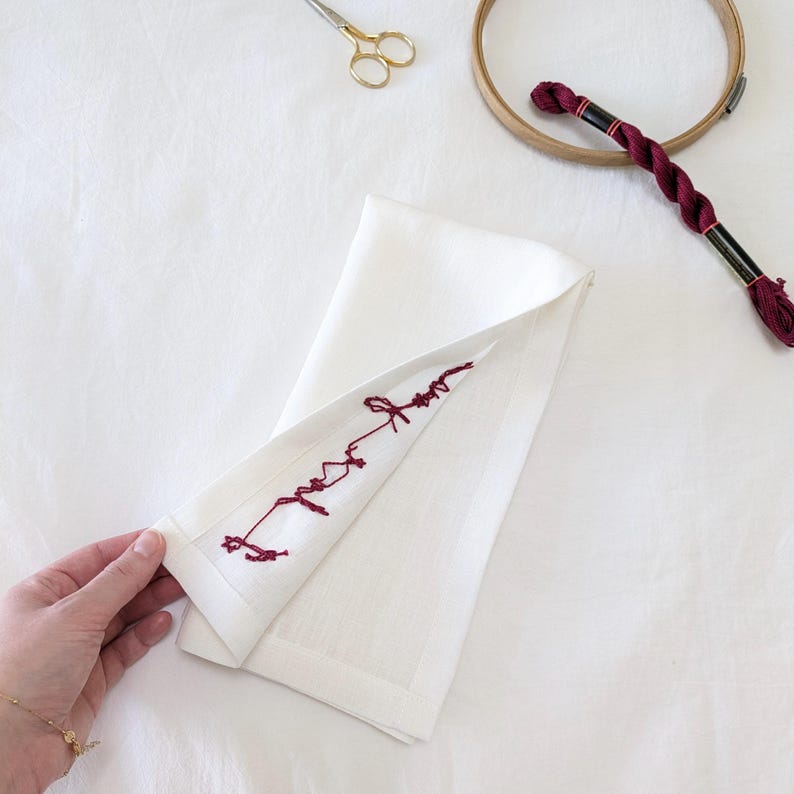 May include: A white linen napkin with a red embroidered design. The embroidery is a simple, free-flowing design that looks like a handwritten word.