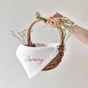 Op de afbeelding: Een rieten mand met een witte linnen voering en de naam "Sammy" geborduurd in roze. De mand is versierd met witte gipskruidbloemen.