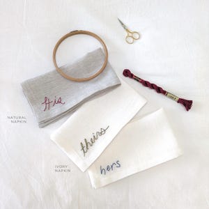 May include: Two embroidered linen napkins, one natural and one ivory, with the words "his", "theirs", and "hers" stitched in different colors. A spool of embroidery floss and a pair of scissors are also visible.