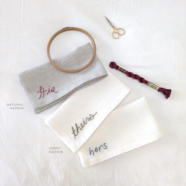 May include: Two embroidered linen napkins, one natural and one ivory, with the words "his", "theirs", and "hers" stitched in different colors. A spool of embroidery floss and a pair of scissors are also visible.