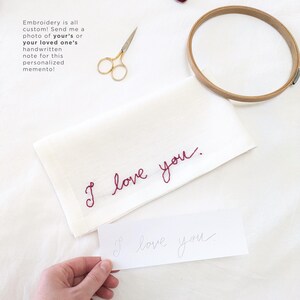 May include: A white linen tea towel with the phrase "I love you" embroidered in red thread. A handwritten note with the same phrase is shown in the foreground.