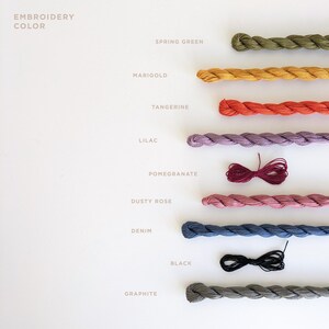 May include: A collection of embroidery floss spools in various colors, including spring green, marigold, tangerine, lilac, pomegranate, dusty rose, denim, black, and graphite. The floss is arranged in a vertical line, with the color names written next to each spool.