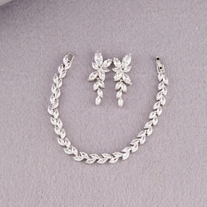 May include: A silver bracelet and earrings set with clear cubic zirconia stones. The bracelet is a delicate chain with leaf-shaped stones. The earrings are dangle earrings with a cluster of stones in the shape of a flower.