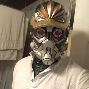 May include: A white shirt with a button-up collar and a silver and gold helmet with red eyes. The helmet has a gold and black design with a gold crown.