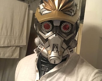 Può includere: Una camicia bianca con colletto abbottonato e un casco argentato e dorato con occhi rossi. Il casco ha un design dorato e nero con una corona dorata.