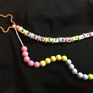 Könnte beinhalten: Eine weiße Schnur mit einem sternförmigen Metallanhänger an einem Ende und Alphabetperlen in einem Regenbogenfarbendesign. Die Schnur hat auch eine separate Strähne aus pastellfarbenen Perlen.