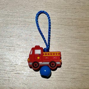 Könnte beinhalten: Ein roter Feuerwehrauto-Charm mit einer blauen Kordelschlaufe und einer blauen Perle. Der Feuerwehrauto-Charm hat gelbe und weiße Details, einschließlich eines Sterns an der Seite. Der Charm ist für einen Schuh bestimmt.
