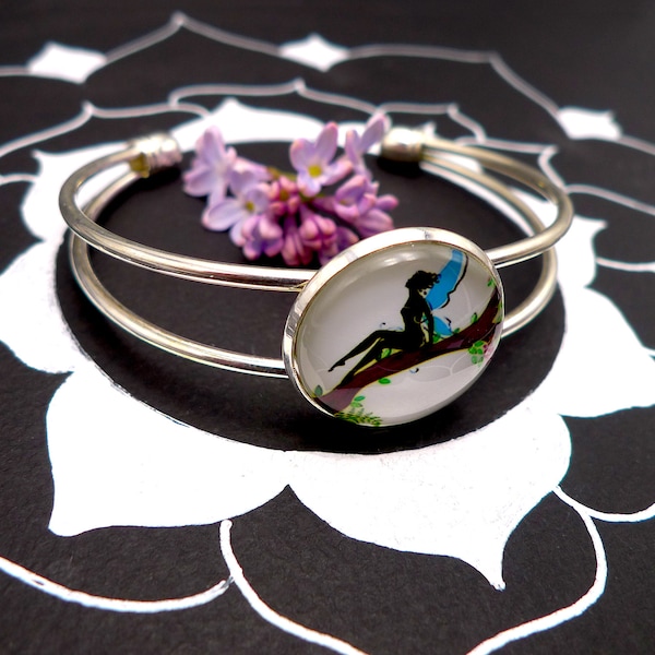 Bracelet jonc en métal argenté clair avec cabochon en verre, fée aux ailes bleues assise sur un tronc d'arbre dans la nature