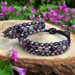 Könnte beinhalten: Zwei handgefertigte Armbänder mit drei Reihen aus lila und hellgrünen Perlen, auf schwarzem Kordelband aufgefädelt. Die Armbänder werden auf einer Holzoberfläche präsentiert, mit einem verschwommenen Hintergrund aus rosa Blüten.