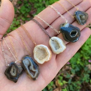 Peut inclure: Une main tenant six colliers avec des pendentifs en cristal d'agate de différentes couleurs. Les pendentifs sont tous en forme de géode, avec un centre creux et un extérieur lisse et poli. Les colliers sont faits de chaînes argentées, dorées et noires.
