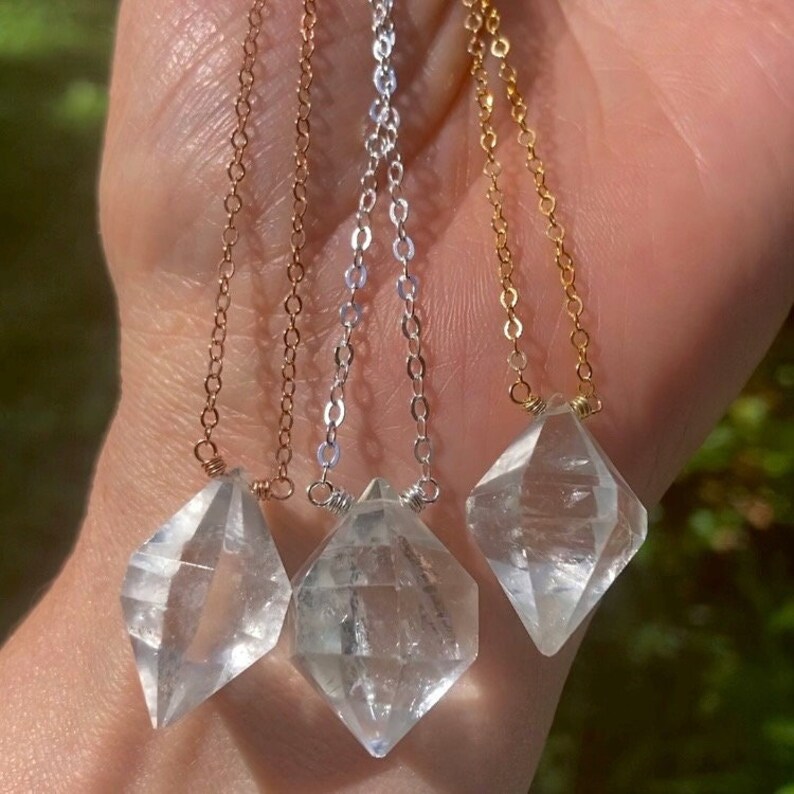 May include: Three necklaces with clear quartz crystal pendants in the shape of a diamond. The necklaces are on a hand and have different chain styles: rose gold, silver, and gold.