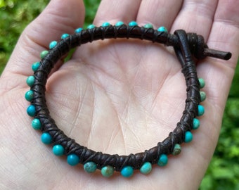 May include: A brown leather bracelet with turquoise beads. The bracelet is wrapped around a hand.