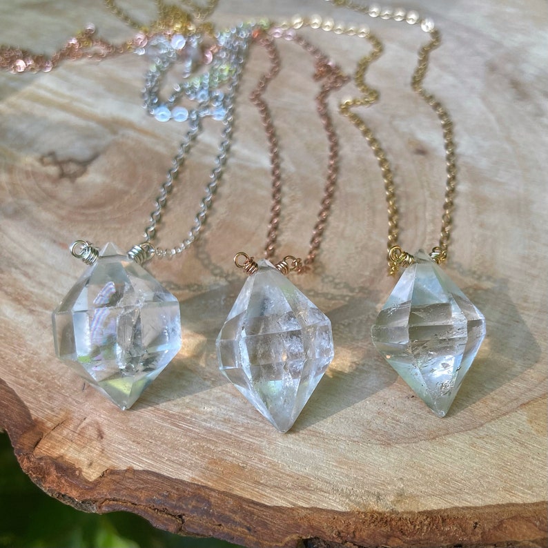 May include: Three necklaces with clear diamond-shaped crystal pendants on a wooden surface. The necklaces are made with different colored chains: silver, rose gold, and gold.