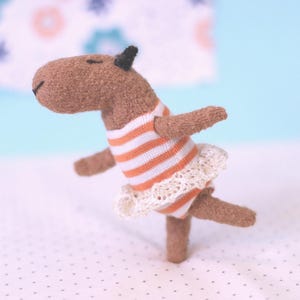 May include: A handmade brown capybara plush toy in a ballet pose. The capybara wears a striped orange and white shirt and a white lace tutu. The toy has small black ears and closed eyes, giving it a peaceful expression.