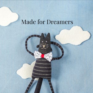 Könnte beinhalten: Ein schwarzes Katzen-Stofftier mit grauen Querstreifen, das eine weiße Fliege mit Punkten und roter Mitte trägt. Die Katze ist vor einem hellblauen Hintergrund mit weißen Filzwolken und dem schwarzen Text Made for Dreamers platziert.