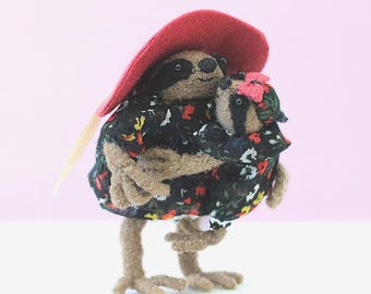 Puede incluir: Escultura de fieltro hecha a mano de una madre y un niño, ambos parecidos a perezosos. Llevan vestidos negros con un estampado floral. La madre perezosa lleva un sombrero rojo grande. El niño tiene un lazo rosa.