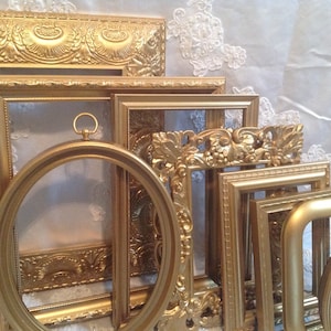 May include: A collection of ornate, gold-colored picture frames in various shapes and sizes. The frames have detailed carvings and embellishments, including floral and scroll designs. One frame is circular, and another has a small ring at the top. The frames are arranged against a white, textured background.