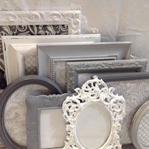 May include: A collection of gray and white picture frames with ornate designs. The frames are arranged in a stack and are all different sizes.