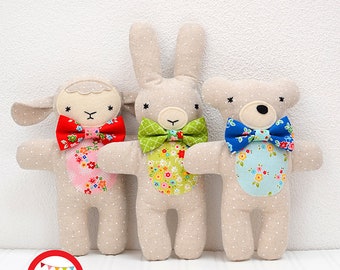 May include: A sewing pattern titled "Three Little Friends" featuring three stuffed animal toys: a lamb, a bunny, and a bear. Each toy has a patterned bow tie and a floral belly patch. The text "SEWING PATTERN" and "NÄHANLEITUNG" are also visible.