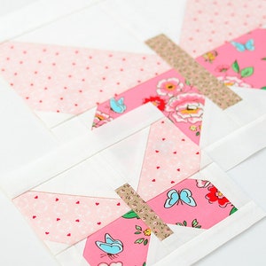 May include: A pink and white butterfly quilt block with a floral pattern and blue butterflies. The block is made of fabric squares sewn together to create a geometric design.