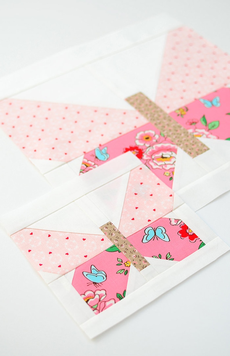 May include: A pink and white butterfly quilt block with a floral pattern and blue butterflies. The block is made of fabric squares sewn together to create a geometric design.