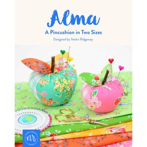 May include: Two apple-shaped fabric pincushions in teal and pink floral patterns with brown stems and heart-topped pins. The text reads Alma A Pincushion in Two Sizes Designed by Nadra Ridgeway. Includes gold scissors and a round pin wheel.