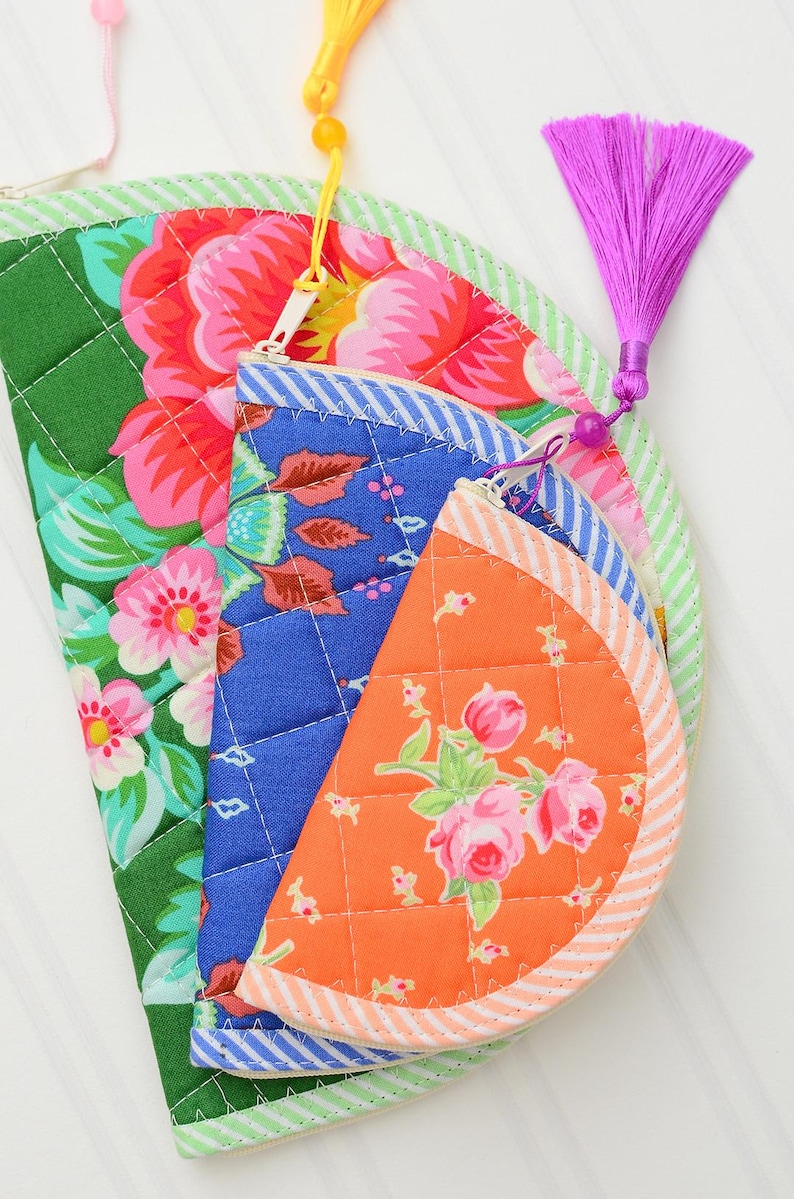 May include: Three semi-circular quilted pouches with zip closures. The largest is green with pink flowers, the middle is blue with a floral pattern, and the smallest is orange with pink roses. Each features a striped border and a colourful tassel.