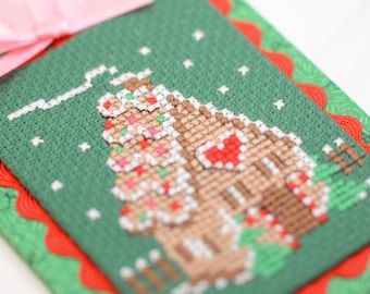 Op de afbeelding: Een rechthoekig kruissteek ornament met een bruin peperkoekhuisje met een rood hart, witte glazuurdetails en groene accenten op een donkergroene stoffen achtergrond. Het stuk is afgezet met rode zigzagband en heeft een lichtroze lintlus.