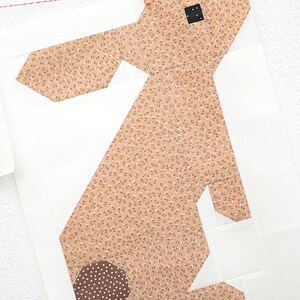 K&ouml;nnte beinhalten: Ein Stoff-Hasen-Quiltblock mit hellbraunem Blumenmuster auf K&ouml;rper und Ohren und einem braunen Tupfenschwanz. Der Hase ist vor einem wei&szlig;en Hintergrund platziert und wird an einer W&auml;scheleine befestigt.