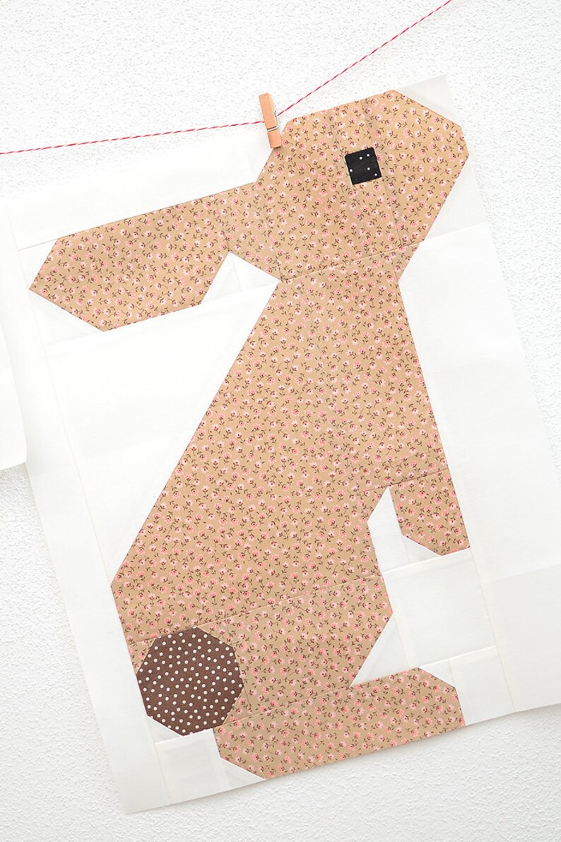 K&ouml;nnte beinhalten: Ein Stoff-Hasen-Quiltblock mit hellbraunem Blumenmuster auf K&ouml;rper und Ohren und einem braunen Tupfenschwanz. Der Hase ist vor einem wei&szlig;en Hintergrund platziert und wird an einer W&auml;scheleine befestigt.