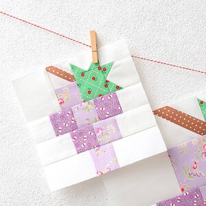 May include: Two unfinished quilt blocks hanging on a clothesline with pegs. The blocks are made of white fabric with a patchwork design of purple, green, and pink floral fabrics. The blocks are hanging on a white wall.