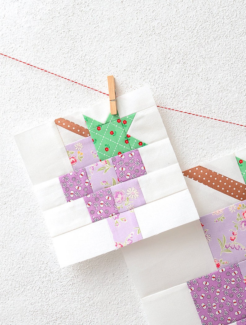 May include: Two unfinished quilt blocks hanging on a clothesline with pegs. The blocks are made of white fabric with a patchwork design of purple, green, and pink floral fabrics. The blocks are hanging on a white wall.
