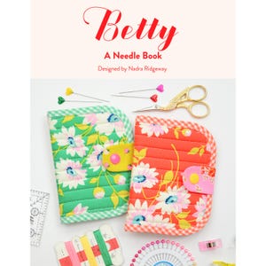Può includere: Un libro per aghi intitolato "Betty" con un design floreale. L'immagine presenta due custodie per aghi in tessuto, una verde e una rossa, con motivi floreali bianchi e rosa. Sono visibili anche accessori per cucire come spilli, forbici e filo.
