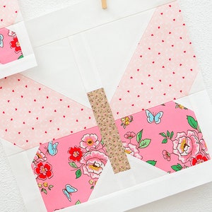 May include: A pink and white butterfly quilt block with a floral pattern and a brown and white stripe in the center. The block is hanging on a white wall with a red string and a wooden clothespin.