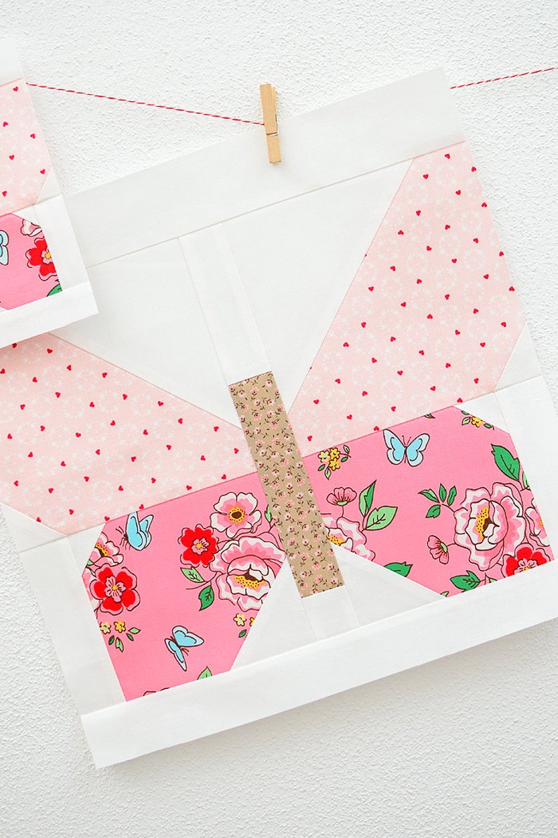 May include: A pink and white butterfly quilt block with a floral pattern and a brown and white stripe in the center. The block is hanging on a white wall with a red string and a wooden clothespin.