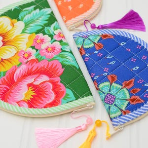 May include: Three semi-circular quilted fabric pouches featuring vibrant floral patterns in green, blue, and orange. Each pouch has a zippered closure with a decorative tassel attached, including pink, yellow, and purple colour options.