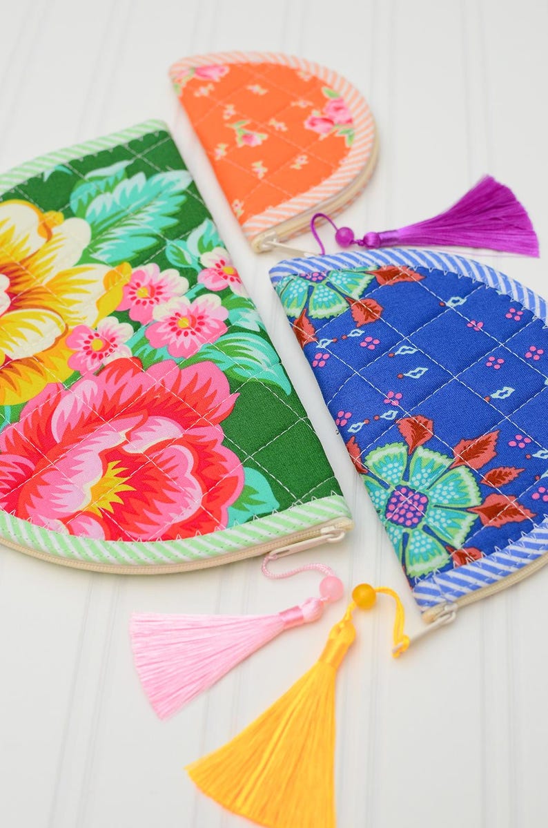 May include: Three semi-circular quilted fabric pouches featuring vibrant floral patterns in green, blue, and orange. Each pouch has a zippered closure with a decorative tassel attached, including pink, yellow, and purple colour options.