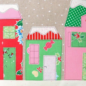 May include: A festive pillow with a snowy scene featuring three houses decorated with red, green, and pink fabrics. The houses have windows, doors, and candy cane accents. The background is a beige fabric with white polka dots.